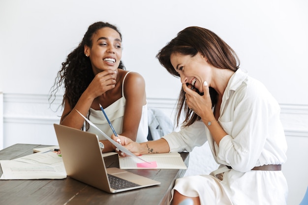 Image of a beautiful young women colleagues at workplace indoors in office work with paper documents and laptop computer.