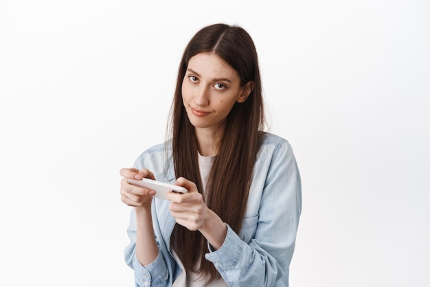 Image of bored and gloomy girl look from under forehead while using her phone playing boring game watching uninteresting video online on smartphone standing over white background