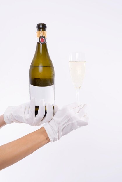 Image of a bottle of champagne and a wine glass in female hands. Light studio background. White gloves. Mixed media