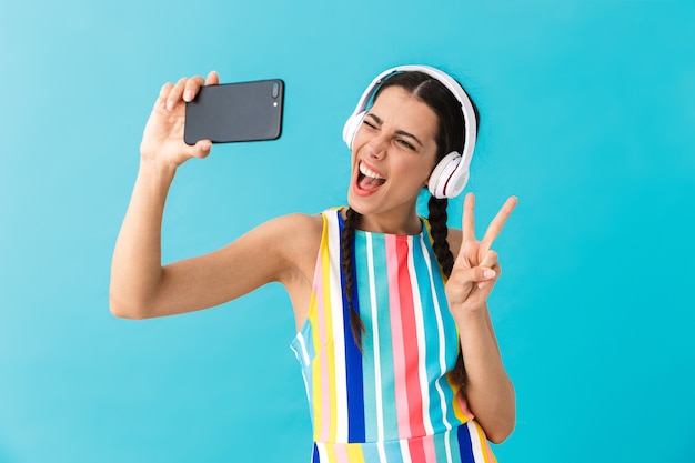 Image of brunette beautiful woman wearing headphones winking while using cellphone isolated over blue wall