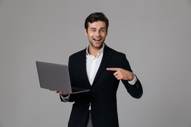 Immagine di uomo d'affari bruna 30s in tuta gioia durante l'utilizzo di laptop, isolato su muro grigio