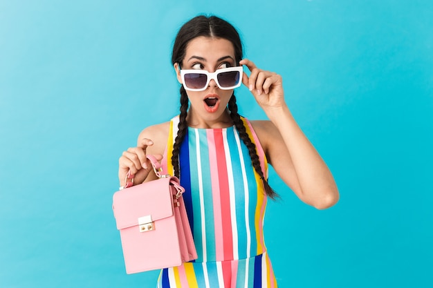 Image of brunette shocked woman wearing sunglasses looking aside while holding pink bag isolated over blue wall