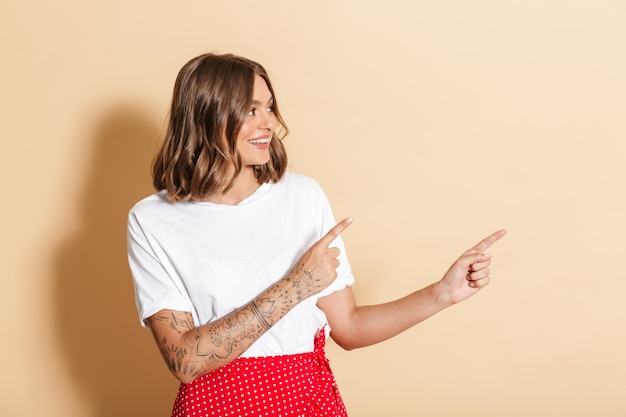 Image of brunette woman in red skirt smiling and pointing finger at copyspace isolated over beige wall