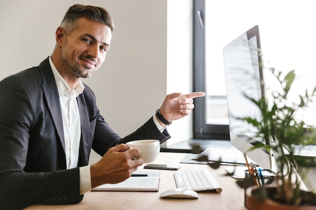 オフィスでコンピューターの作業中にコーヒーを飲むスーツを着ている30代のビジネスライクな男性の画像