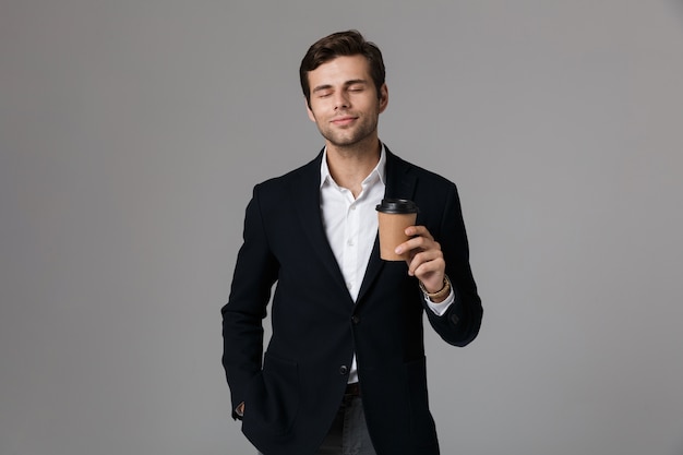 Image of businessman 30s in suit smiling and holding paper cup with coffee, isolated over gray wall