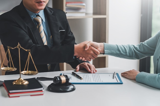 Image of a businessman lawyer shaking hands with a woman client collaboration agreement contract document and a mallet placed at a desk.