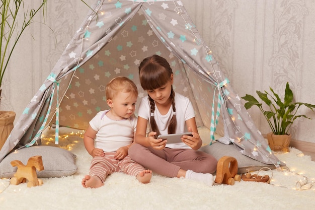 Image of calm little sisters wearing white t shirts sitting in wigwam and watching cartoons together with concentration being interested in a new episod posing shelter at home