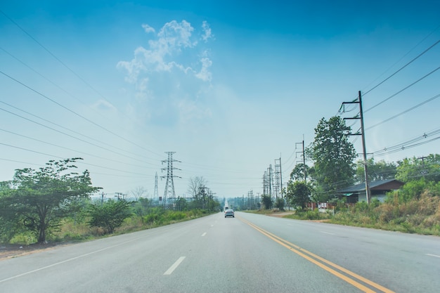 空と雲を背景に、道に沿って木々や街灯柱がある、開いた道路をクルージングしている車の画像。