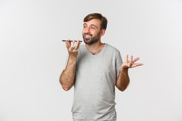Image of carefree guy with beard, wearing basic t-shirt, talking on speakerphone, holding mobile