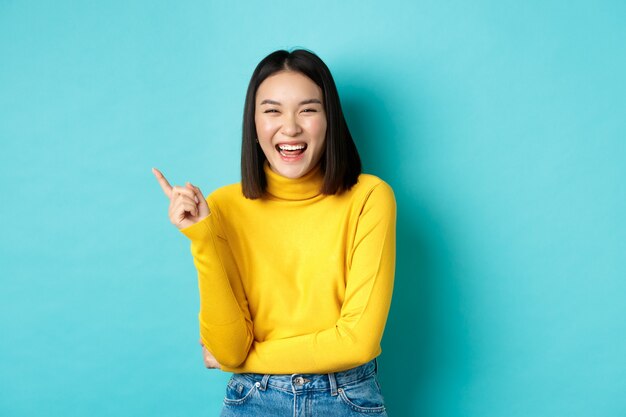 Image of carefree pretty korean girl laughing and looking happy, pointing finger at upper left corner promo, standing against blue background