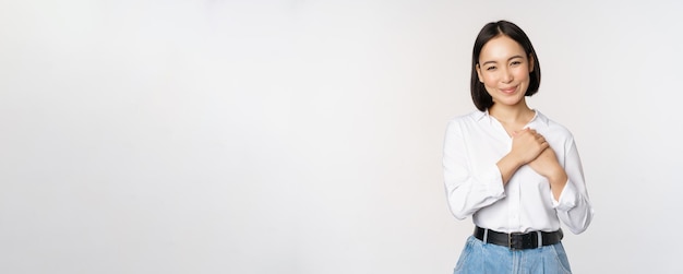 Image of caring young beautiful asian woman holding hands on heart looking pleased and happy smiling at camera standing over white background