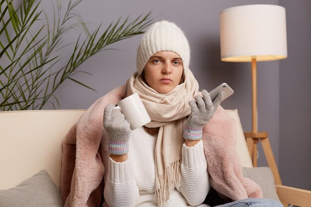 Image of Caucasian attractive woman sitting on the sofa at home wearing winter coat hat and mittens holding smart phone in hands looking at camera posing with cup in hands drinking hot tea