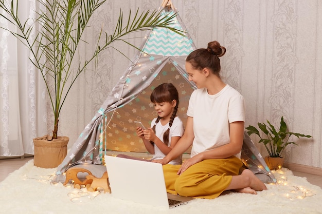 Image of Caucasian dark haired woman sitting on floor near children's teepee with her daughter little girl using mobile phone watching cartoons family use laptop