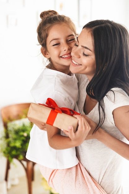 Image of caucasian family brunette mother smiling and giving present box to her little daughter while celebrating birthday at home