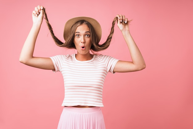 Image of caucasian happy woman wearing straw hat making fun with her hair and grimacing isolated over pink wall