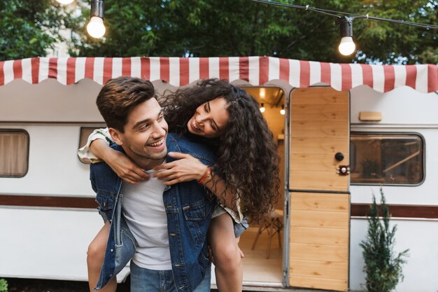 Image of caucasian man wearing denim clothes giving piggyback ride to pretty woman near house on wheels outdoors