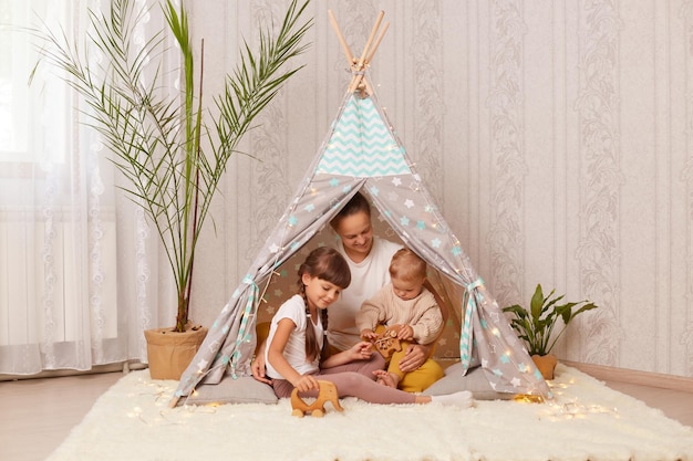 Image of Caucasian mother with her children sitting together teepee tent mom playing with kids in play tent mommy spending weekend with kids playing and having fun in wigwam