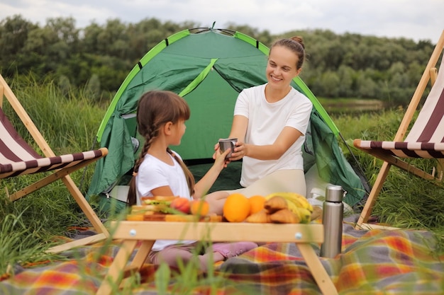 Image of charming delighted young adult woman and little girl sitting on blanket near tent having picnic daughter giving a cup of tea or coffee to her mommy