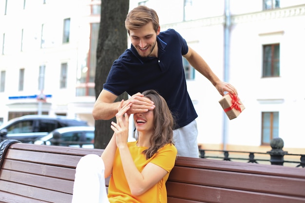 Image of charming excited couple in summer clothes smiling and holding present box together while sitting on bench.