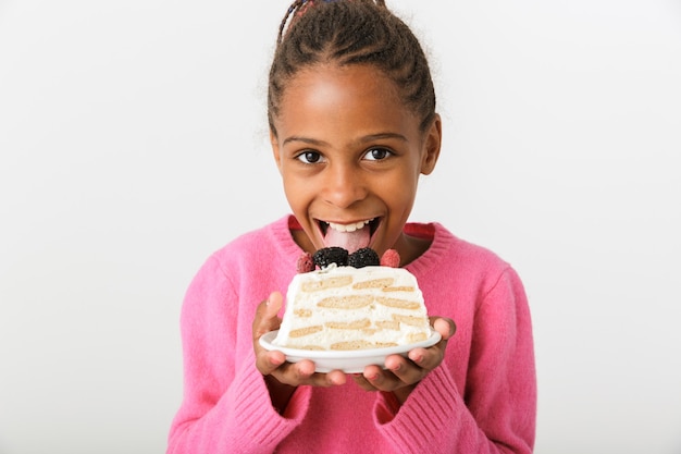 Image of cheerful african american girl eating piece of torte while looking at camera isolated over white wall