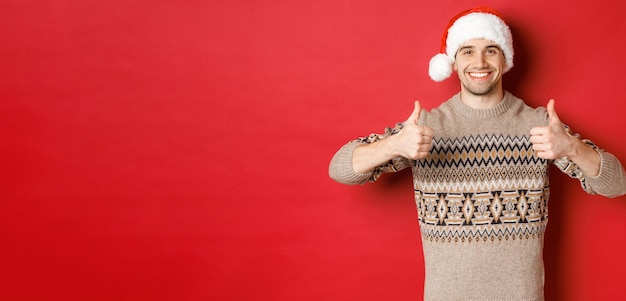 Image of cheerful attractive man in winter sweater and santa hat showing thumbsup celebrating christ...