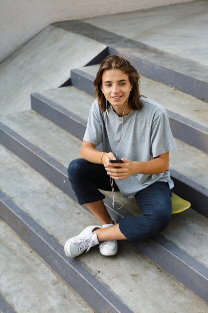 Photo image of cheerful attractive young skater guy sit in the park listening music with earphones using phone.