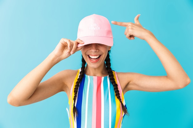 Image of cheerful brunette girl wearing summer dress and cap smiling at camera isolated over blue wall