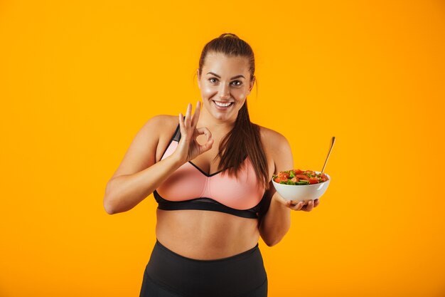 Image of cheerful chubby woman in tracksuit holding plate with green salad, isolated over yellow background