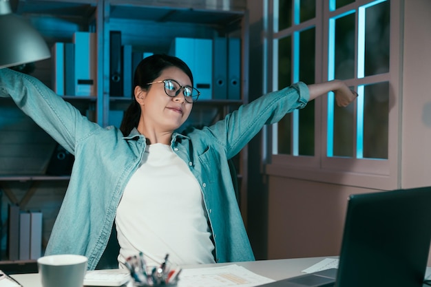 Image of cheerful female designer working late at night with
computer and stretching arms in home office. young worker in
glasses satisfied smiling looking at laptop computer screen and
raised hands