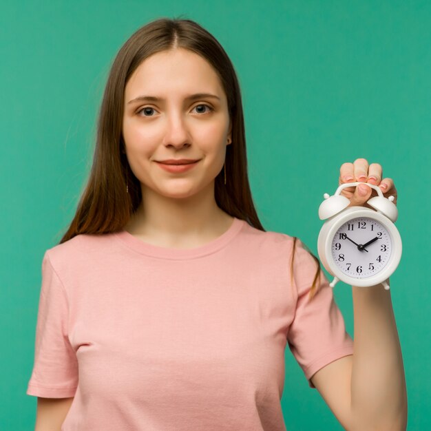 Image of cheerful female student standing over blue. Looking camera with clock alarm in hand.