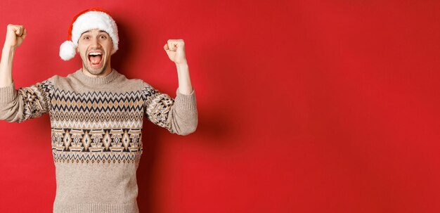 Image of cheerful handsome man in swearer and santa hat celebrating new year triumphing or winning s