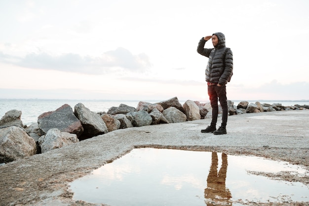 Image of cheerful man dressed in warm clothes walking on the beach. Looking at beach.