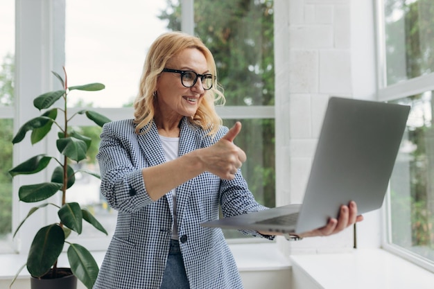 Image of cheerful mature businesswoman standing at office using laptop computer Portrait of a smiling senior lady holding laptop computer