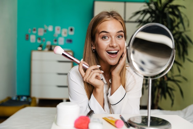 Image of a cheerful smiling young cute girl in pajama at home near mirror with makeup brush.