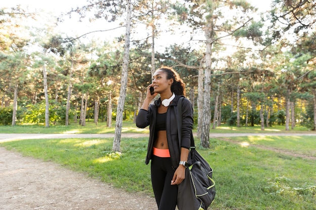 Image of cheerful woman 20s wearing black tracksuit and headphones, speaking on mobile phone while walking through green park