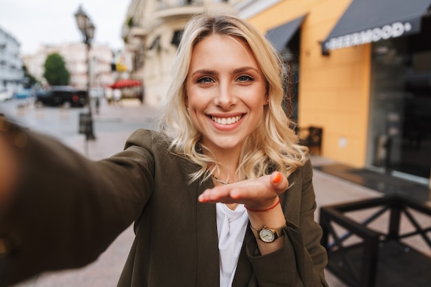 Image of cheerful woman smiling and taking selfie photo, while walking through city street