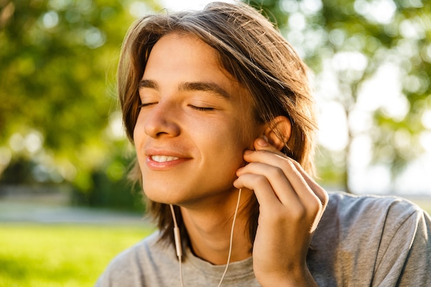 Image of cheerful young guy listening music with earphones in the park.