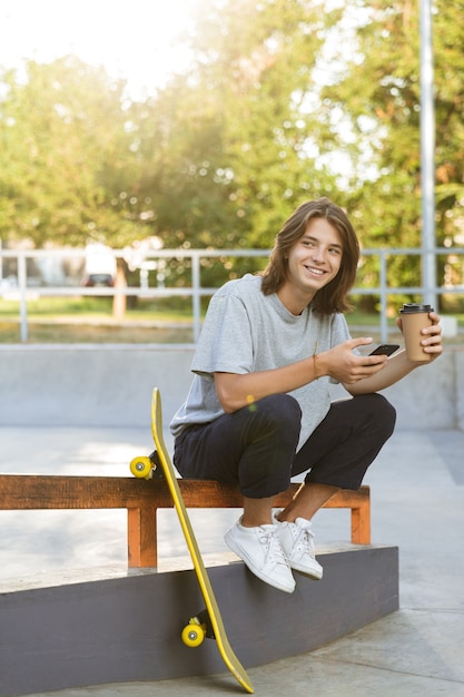 Image of cheerful young skater guy sit in the park with skateboard using mobile phone drinking coffee.