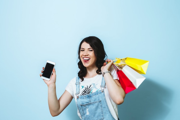 Image of a cheerful young woman showing blank screen mobile phone while carrying isolated over yellow wall shopping bags