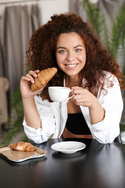 Immagine di una giovane donna piuttosto ottimista allegra vestita con una camicia bianca al chiuso in un hotel di casa in cucina che beve caffè mangia croissant fa colazione.