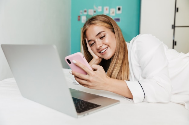 ラップトップコンピューターと携帯電話を使用して、自宅のベッドでパジャマを着た陽気な若いかわいい女の子の画像。