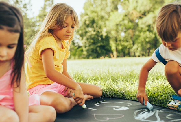 Image of children sitting on the green grass playing with colorful chalks Happy little kids drawing with chalks in the park Friends boy and two girls having fun on sunlight outdoors Childhood