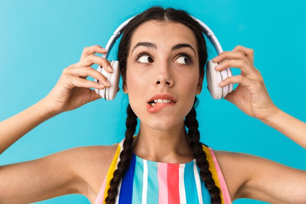 Image closeup of brunette puzzled woman looking aside and biting her lip while using headphones isolated over blue wall