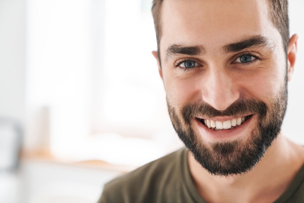 Foto primo piano dell'immagine di un uomo caucasico felice che indossa una maglietta casual che guarda e sorride alla telecamera mentre lavora in un ufficio luminoso