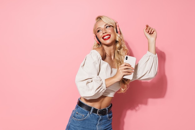 Image closeup of cheerful seductive woman wearing red lipstick listening to music with wireless headphones and cellphone isolated over pink wall