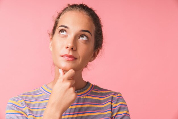 Image closeup of dreamy beautiful woman dressed in colorful clothes thinking and looking upward at copyspace isolated over pink wall