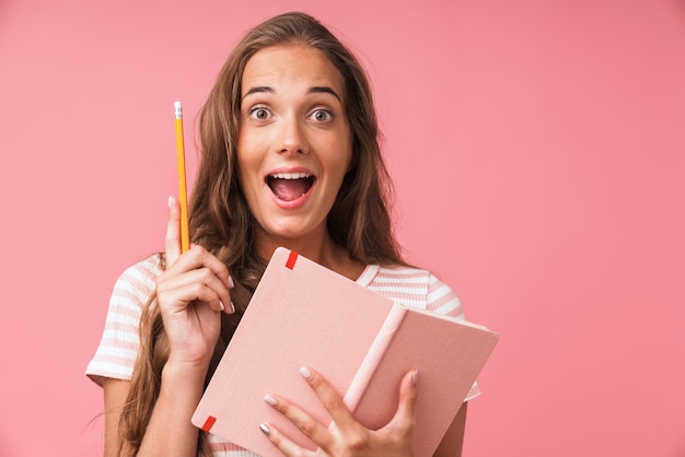 Image closeup of happy young woman wearing striped t-shirt looking while holding diary and pencil isolated over pink wall