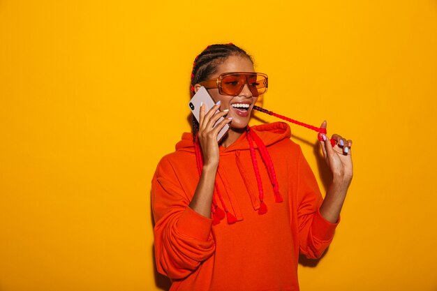 Image closeup of nice african american woman wearing sunglasses and hoodie smiling while talking on cellphone isolated over yellow