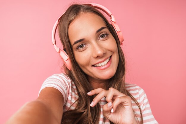 분홍색 벽에 격리된 헤드폰으로 웃고 음악을 들으며 줄무늬 옷을 입은 긍정적인 아름다운 소녀의 이미지 클로즈업