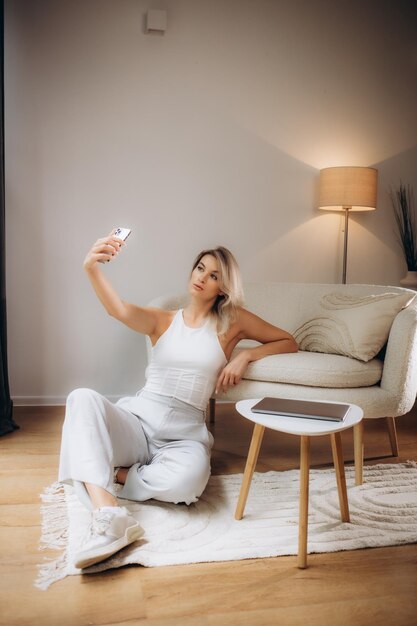 Image closeup of pretty blond woman 20s wearing white tshirt smiling while looking at camera and taking selfie photo in living room